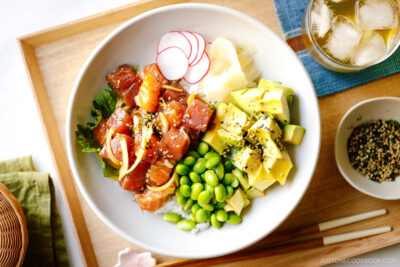 A bowl containing ahi tuna and salmon poke along with edamame, avocado, sushi ginger, and radish over bed of steamed rice.